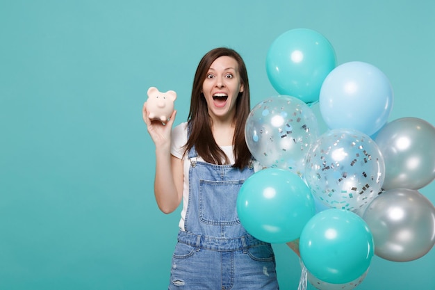 Surprised young woman keeping mouth wide open, holding piggy money bank, celebrating with colorful air balloons isolated on blue turquoise background. Birthday holiday party, people emotions concept.
