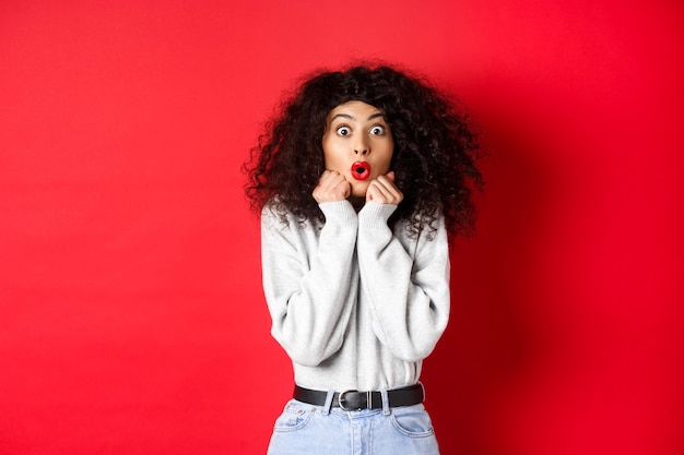 Surprised young woman looking with disbelief and amazement , saying wow and standing in awe on red wall