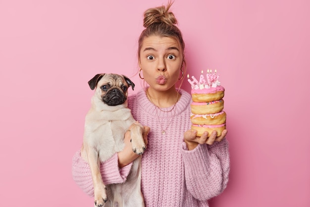 Surprised young woman pet owner keeps lips folded looks with wonder at camera holds birthday doughnuts with candles and pug dog celebrates bday of favorite pet poses against rosy background