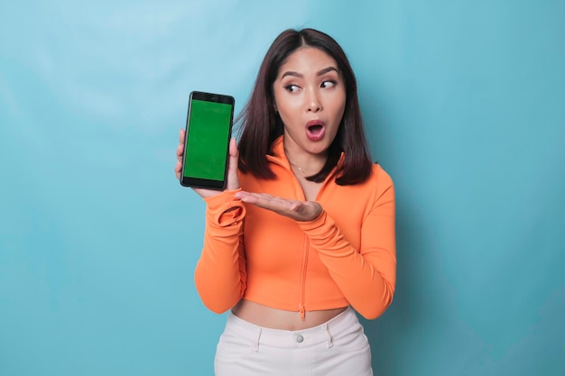 A surprised young woman pointing at copy space on her smartphone in her hand isolated on blue background