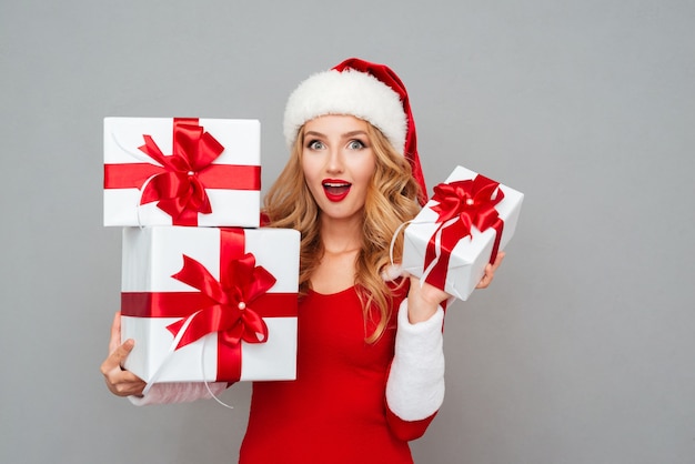 Surprised young woman in red santa claus outfit holding heap of gift boxes isolated on the gray surface