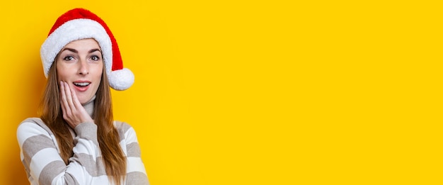 Surprised young woman in Santa Claus hat on a yellow background. Banner.