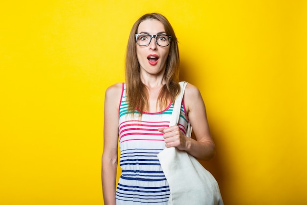 Surprised young woman in striped dress, glasses and with linen bag on yellow background.