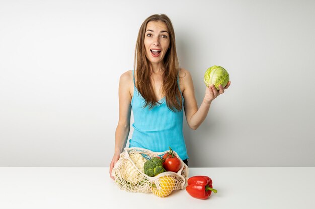 Surprised young woman taking out vegetables from bag on table on white background