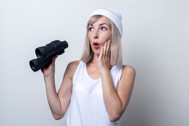 Surprised young woman in a white hat with binoculars on a light background.