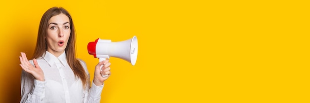 Surprised young woman in a white shirt holds a megaphone on a yellow background Hiring concept ad