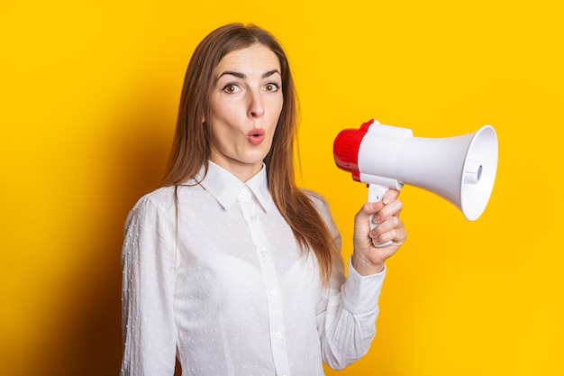 Surprised young woman in a white shirt holds a megaphone on a yellow background