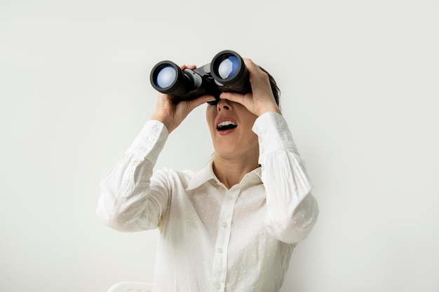 Surprised young woman in a white shirt looks through binoculars on a white background