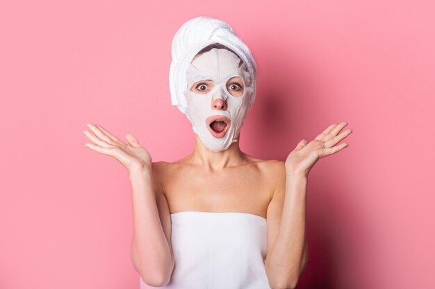 Surprised young woman with a cosmetic mask on her face on a pink background, skin care