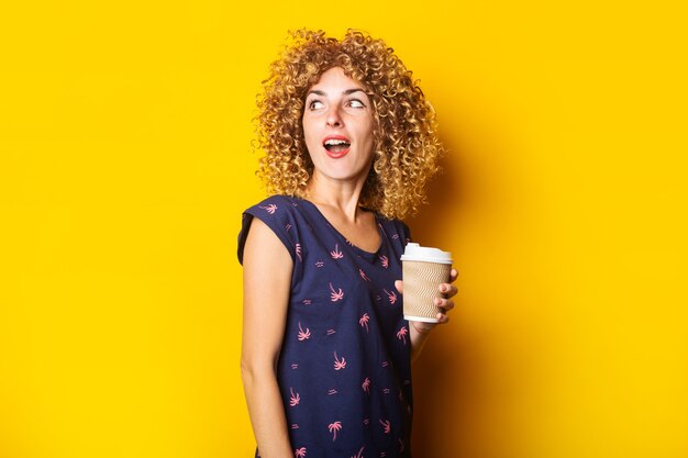 Surprised young woman with curly hair holding paper cup on yellow surface