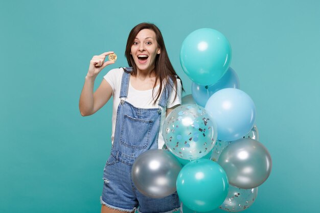 Surprised young woman with opened mouth holding bitcoin, future currency, celebrating with colorful air balloons isolated on blue turquoise background. Birthday holiday party, people emotions concept.