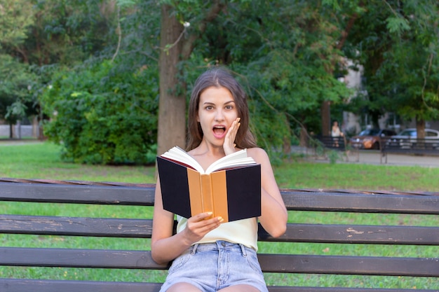 입을 크게 벌리고 뺨에 손을 대고 놀란 젊은 여성이 야외에서 책을 읽고 있습니다.