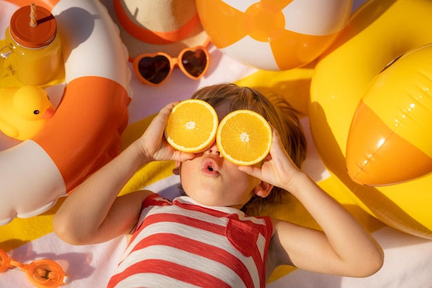 Surprized child holding slices of orange fruit like sunglasses\
kid wearing striped yellow tshirt lying on beach towel healthy\
eating and summer vacation concept