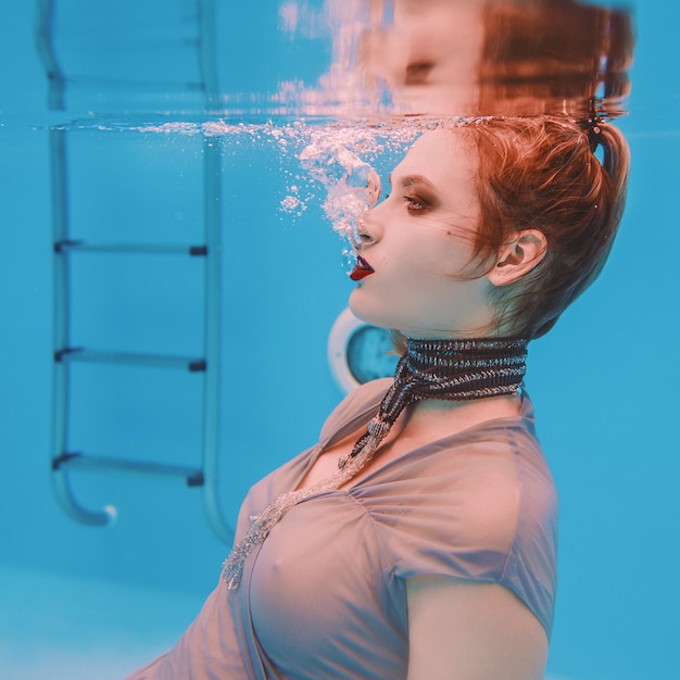 surreal art portrait of young woman in grey dress and beaded scarf underwater in the swimming pool