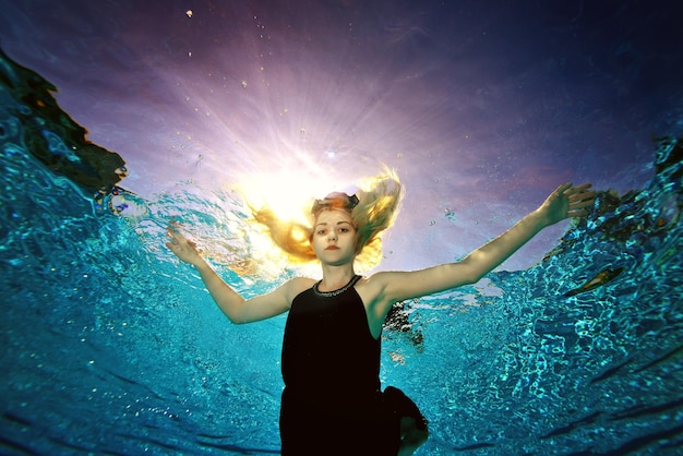 Surreal portrait of a blonde girl posing underwater in a black dress with her arms outstretched