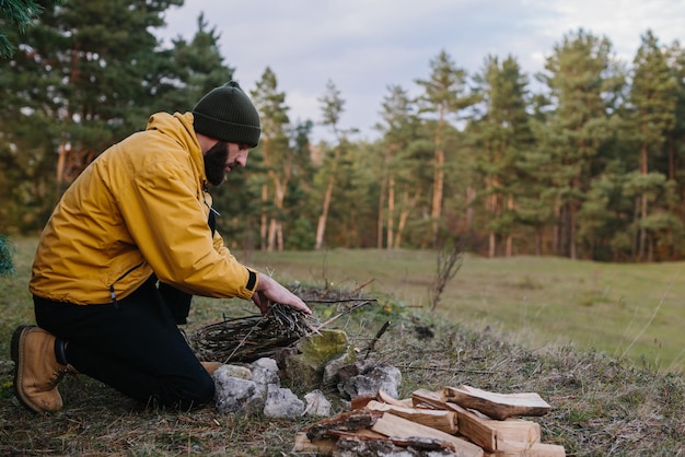 Выживание в дикой природе Бородатый мужчина разжигает костер возле импровизированного укрытия из сосновых веток
