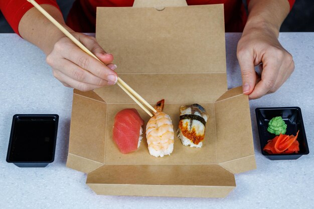 Sushi met tonijn garnalen en paling ligt in een kartonnen doos op tafel in de keuken Het meisje in de rode trui eet met stokjes Fastfood concept