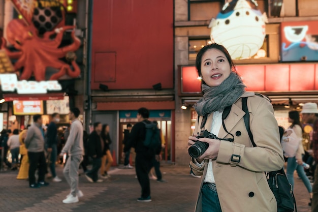 sushi-restaurant met Fugu pufferfish-ballonstandbeeld en takoyaki-octopusteken op de achtergrond. jong meisje dat vrolijk camera vasthoudt terwijl ze 's nachts in de winkelstraat Namba van Dotonbori staat.