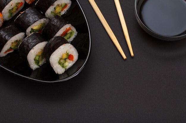 Sushi rolls in nori seaweed sheets with avocado and red fish on ceramic plate. Bowl with soy sauce and wooden sticks. Top view with black background.