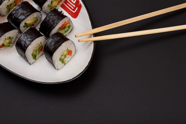 Sushi rolls in nori seaweed sheets with avocado and red fish on ceramic plate and wooden sticks. Top view with black background and copy space.