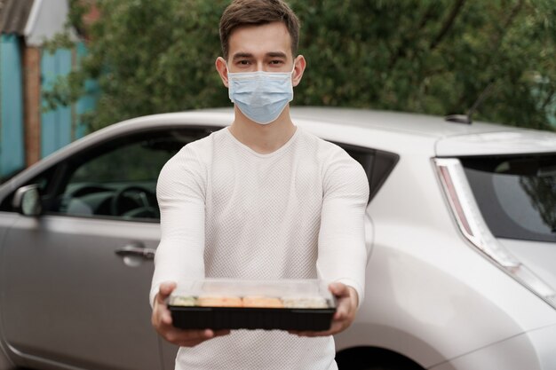Sushi set in box healthy food delivery online service by car. Handsome man courier in medical mask gives sushi box to you. Japanese cuisine.