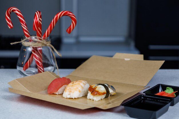 Sushi with tuna shrimp and eel lies in a cardboard box on the table in the kitchen Christmas concept Festive decorations in the form of candles figurines and sweets