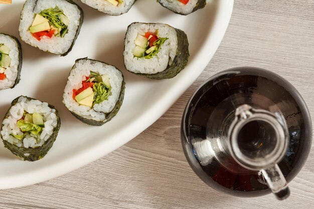 Sushibroodjes met rijst, stukjes avocado, komkommer, rode paprika en slablaadjes op keramische plaat, eetstokjes, glazen fles met sojasaus. Bovenaanzicht