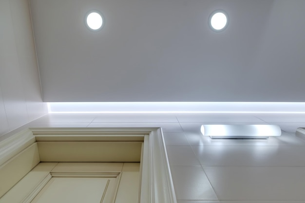 Подвесной потолок с галогенными точечными лампами и гипсокартонной конструкцией в пустой комнате в квартире, офисе, клинике, отеле или доме. Натяжной потолок белого цвета и сложной формы.