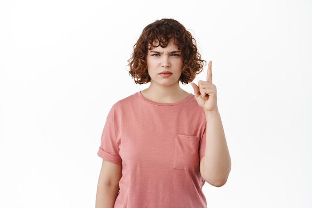 Suspicious curly girl, frowning disappointed and angry, pointing finger up at something bad, standing displeased and bothered in t-shirt, white background.