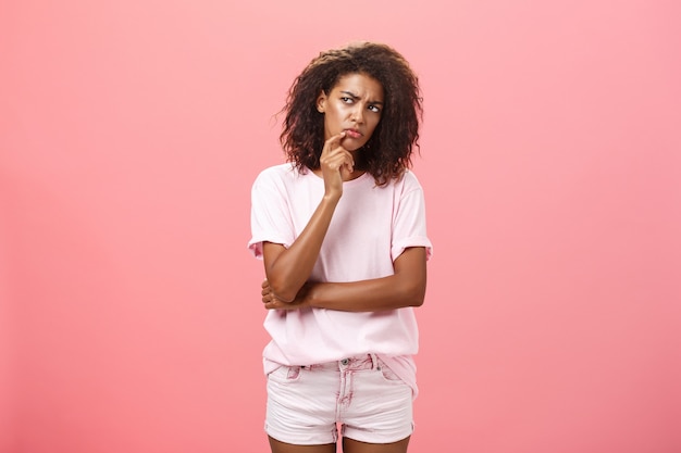 Suspicious dark skinned young curly-haired woman in tshirt touching chin and staring left doubtfully