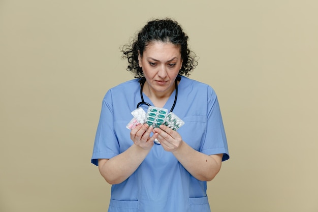 Sospettosa dottoressa di mezza età che indossa uniforme e stetoscopio intorno al collo che tiene confezioni di pillole con entrambe le mani che le guardano isolate su sfondo verde oliva