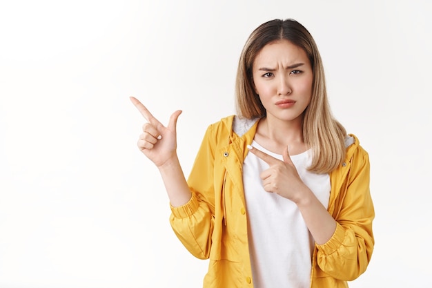 Sospetto insicura ragazza bionda asiatica accigliata socchiudendo gli occhi dubbioso indicando l'angolo in alto a sinistra guarda la macchina fotografica insicura intensa espressione esitante incerta sullo strano prodotto, in piedi muro bianco