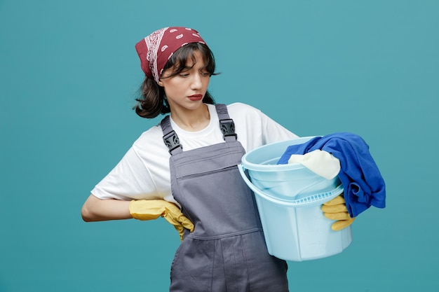 Suspicious young female cleaner wearing uniform bandana and rubber gloves holding and looking at bucket of dirty laundry while keeping hand on waist isolated on blue background