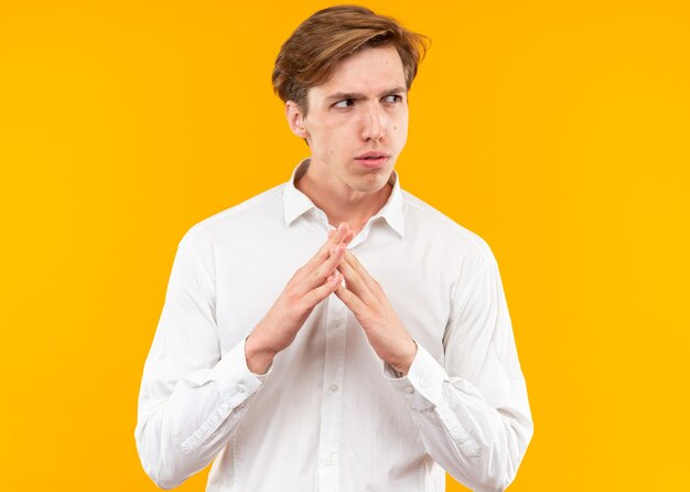 Suspicious young handsome guy wearing white shirt holding hands together isolated on orange wall