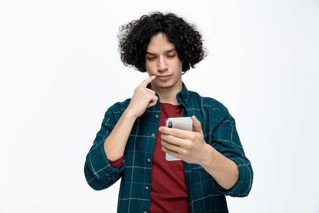 Suspicious young handsome man holding and looking at mobile phone keeping finger on lip isolated on white background