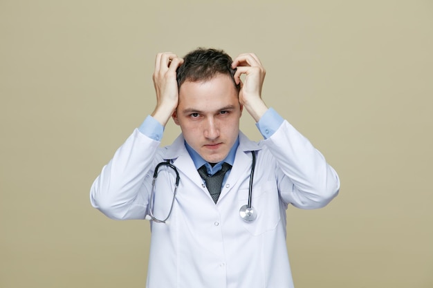 suspicious young male doctor wearing medical robe and stethoscope around neck looking at camera while keeping hands on head isolated on olive green background