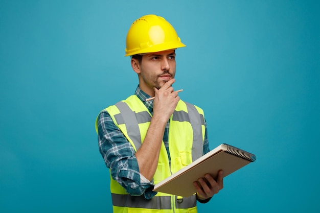 Suspicious young male engineer wearing uniform and safety helmet holding note pad and pencil keeping hand on chin looking at side isolated on blue background