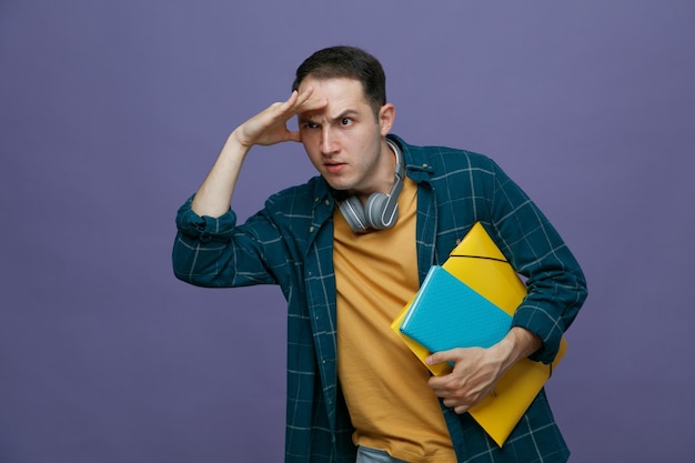 Suspicious young male student wearing headphones around neck holding folder note book keeping hand on forehead looking at side into distance looking for someone isolated on purple background