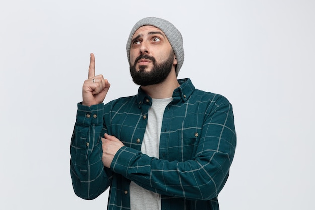 Suspicious young man wearing winter hat looking and pointing up isolated on white background