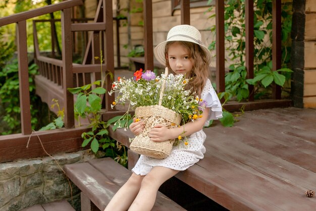 Sute modest little girl with wavy hair in straw bonnet and white dress sitting outdoors on threshold of country house holding wicker basket of wildflowers with both hands Happy summer vacation