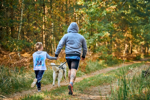 Svetly, Kaliningrad oblast, Russia - October 2, 2021 - Canicross cross country running with dog, father and little son running with Siberian Husky dog, sled dog racing sports competition, back view