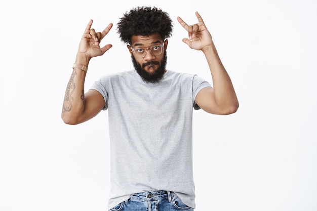 Swah guys. Charismatic and energized cool african american man with beard and tattoos sucking lip raising eyebrows and showing rock-n-roll gesture, standing awesome and chill over gray wall