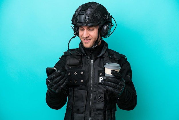 SWAT caucasian man isolated on blue background holding coffee to take away and a mobile