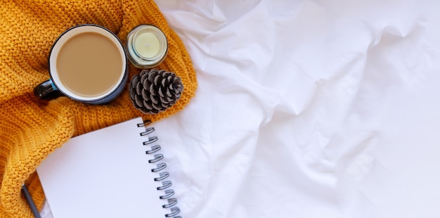 Sweater and cup of coffee with notebook, candles, cone and knitting yeallow clothes on a white crumpled sheet background . Cozy autumn or winter concept.