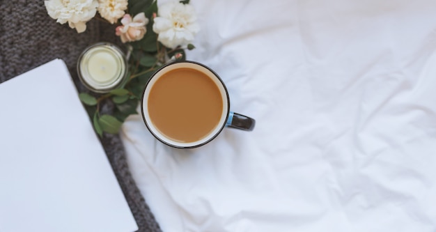 Sweater and cup of coffee with notebook, candles, roses and knitting grey clothes on a white crumpled sheet background . Cozy autumn or winter concept. copy space