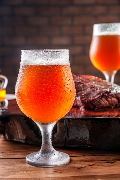 Photo sweaty cold tulipa glasses of ale draft beer with grilled denver steak on wooden cutting board - closeup.