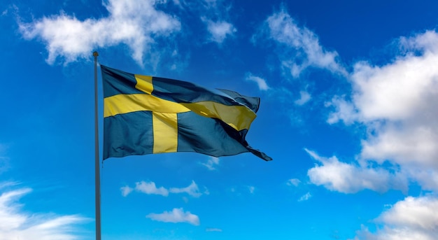 Национальный флаг Швеции развевается на флагштоке голубого облачного неба