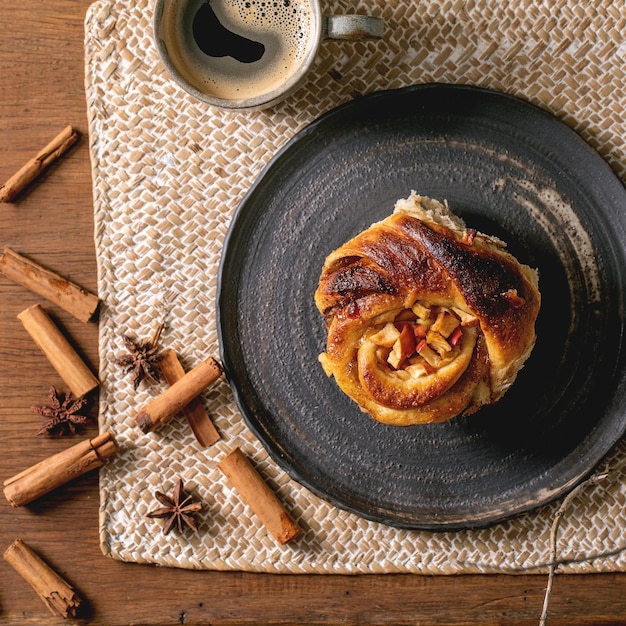 Sweet apple sticky cinnamon roll bun with coffee mug standing with cinnamon sticks and dry branch on straw napkin over wooden background Cozy home bakery Flat lay Square image