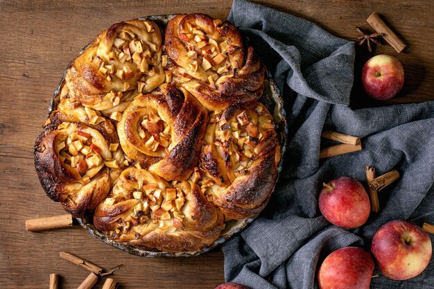달콤한 사과 끈적끈적한 계피 롤빵은 나무 배경에 정원용 사과와 계피 스틱이 있는 둥근 베이킹 트레이에 있습니다. 전통 가정 빵집. 플랫 레이