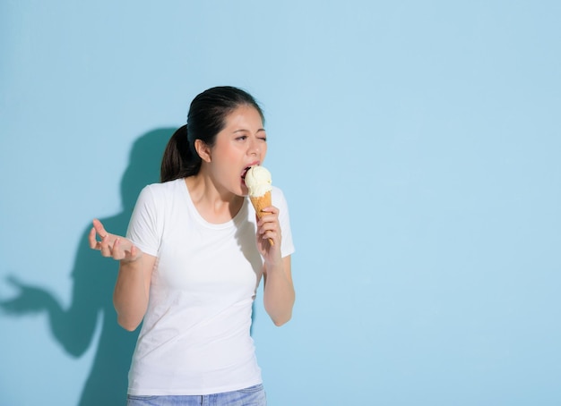 甘い魅力的な女の子は大きな口を開けてバニラアイスクリームを食べ、青い壁の背景に敏感な歯の痛みを伴うパフォーマンスを感じるグラブジェスチャーを作ります.